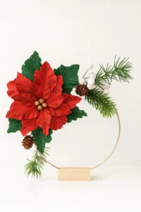 Håndlavet juledekoration med papir-julestjerne i crepepapir, grønne blade, kogler og vintergrene på en metalring med træfod, egnet til både vægophæng og stående dekoration.