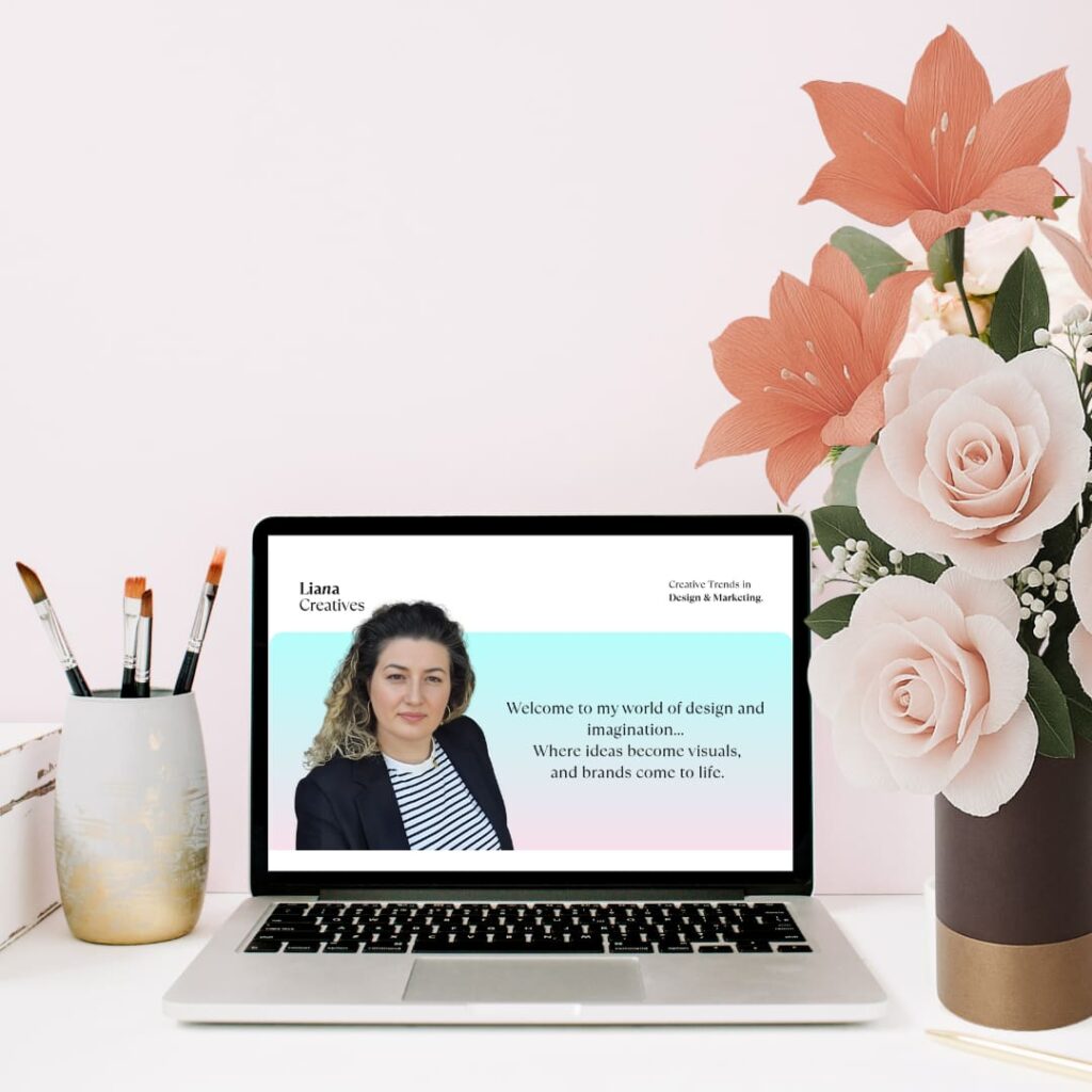 Laptop med Liana Creatives’ designside omgivet af papirroser og koralfarvede papirblomster.